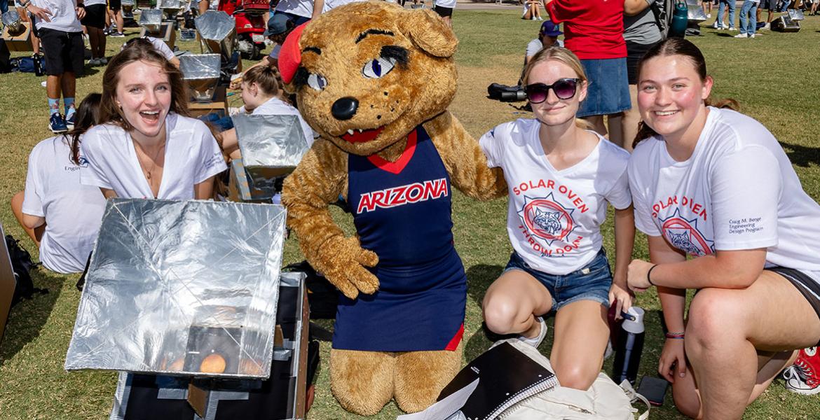 Three students and the Wilma Wildcat mascot pose outdoors with a solar oven