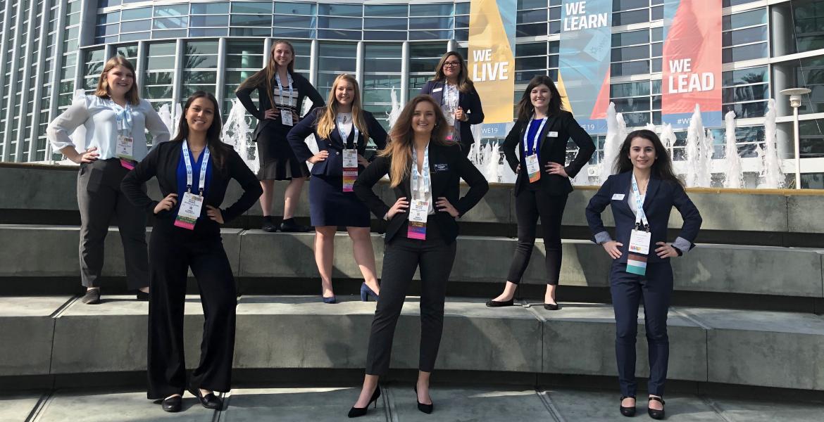 Eight women in professional attire stand on three levels of steps posing for a photo in front of the Anaheim Convention Center.