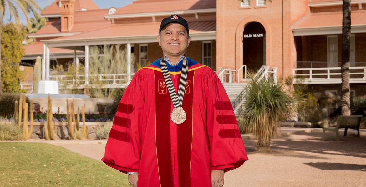 Ricardo Valerdi, wearing academic regalia and a medal, poses in front of Old Main on the University of Arizona campus 