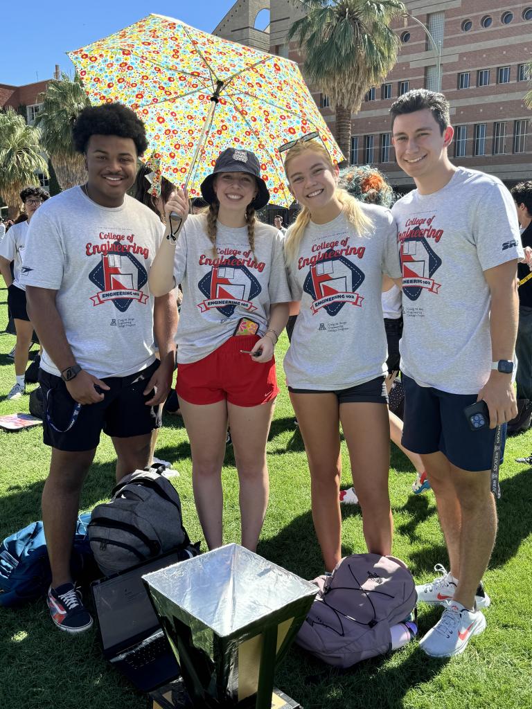 Four students with white shirts take a photo on the University of Arizona grass lawn.