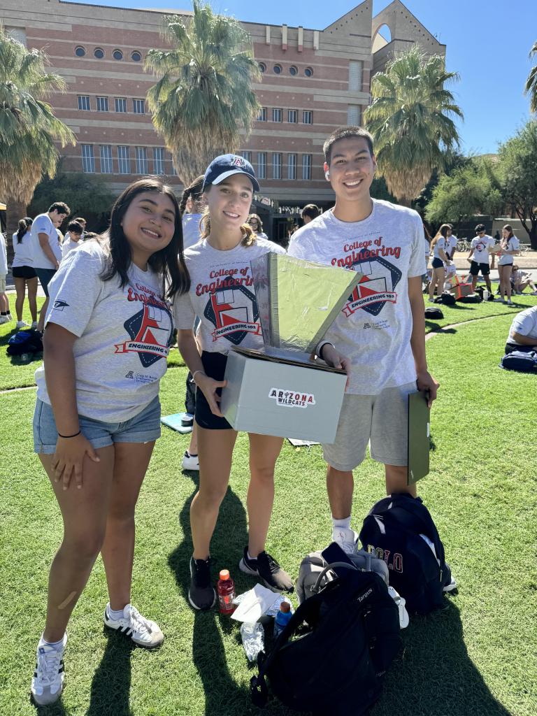 Three students with white shirts take a photo on the University of Arizona grass lawn.