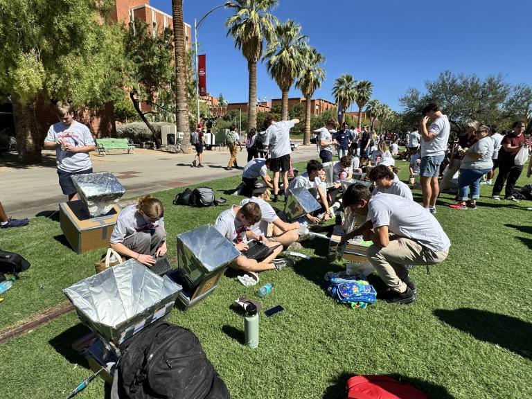 High school students all stand on a grassy lawn and watch their handmade ovens convert the sun's rays into heat.