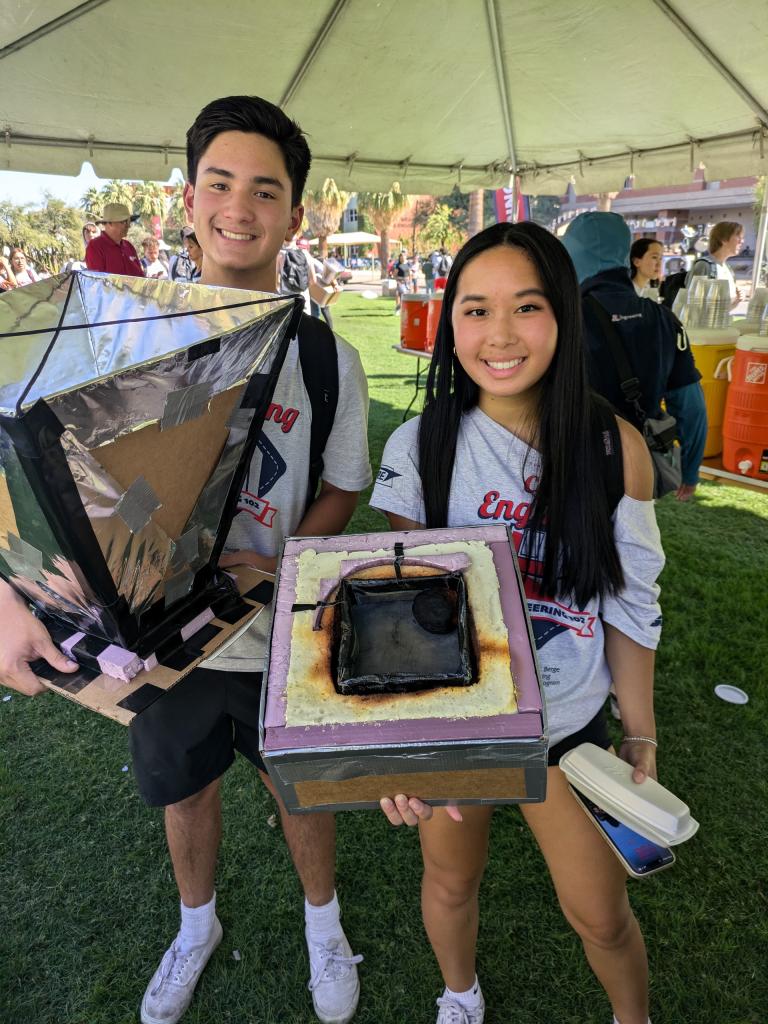 A man and woman with brown hair, Team Solar Nation, hold their handmade oven.