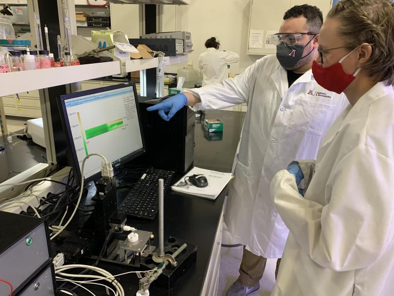 two students wearing lab coats in masks in a laboratory. One is pointing out something on a computer screen to the other.