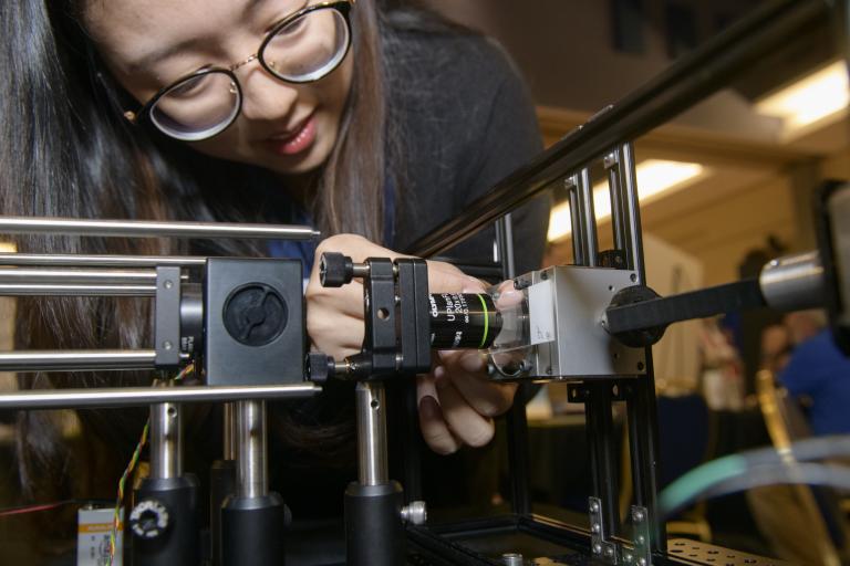 a young woman with glasses leans over to adjust a piece of optical equipment.