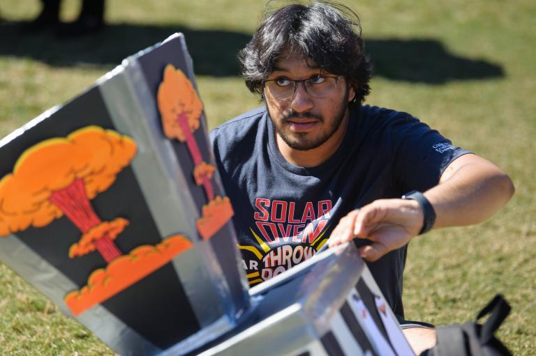 A student looks to the side at his solar oven, a cardboard box decorated with grey duct tape and a red paper cutout of an explosion.