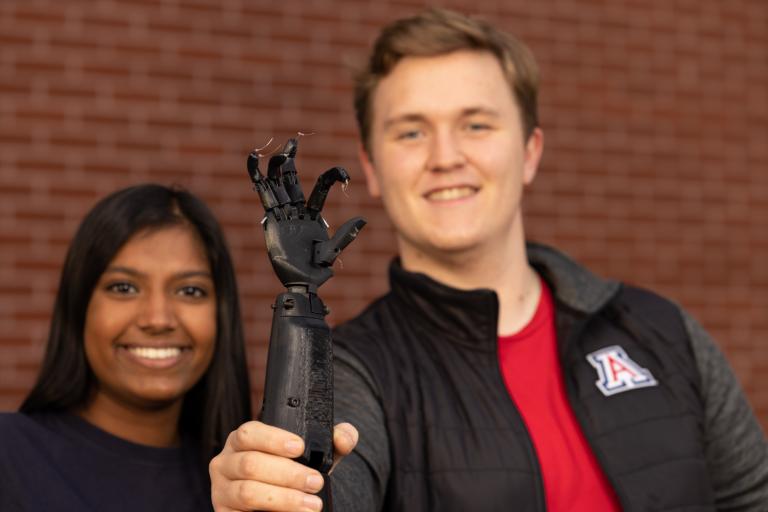 a young man and a woman are smiling, slightly blurred out. In the foreground is a black neuroprosthetic arm, which the man is holding proudly up to the camera.