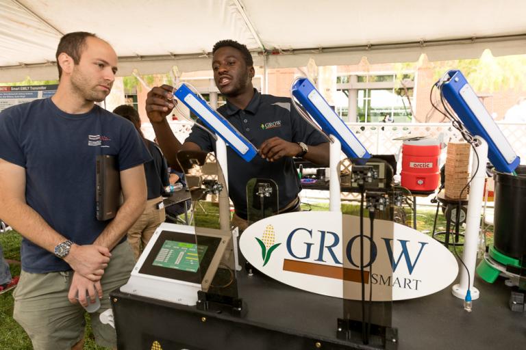 One young man leans over to show another n electronic device. A sign on the machine says "Grow Smart."