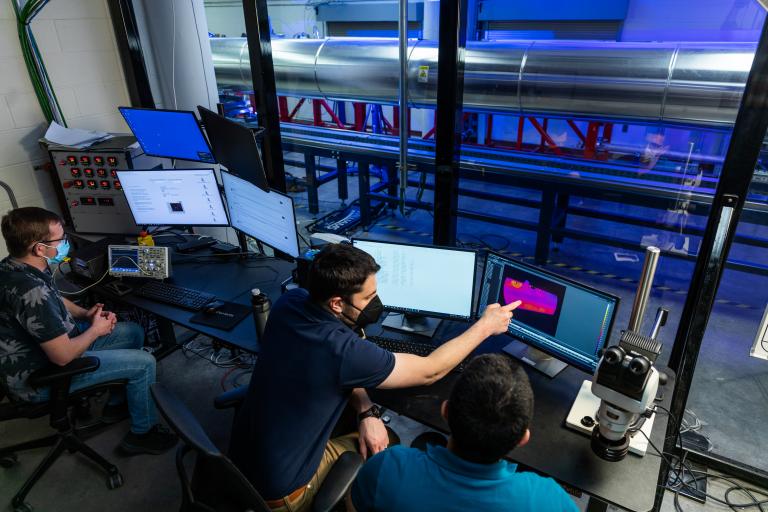 In a room outside a wind tunnel lab, bathed in blue light, a professor points to an image on a screen in front of a student.