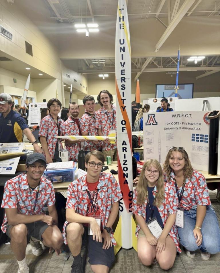 A groupd of eight students in red and white shirts smile at the camera next to their award winning rocket.