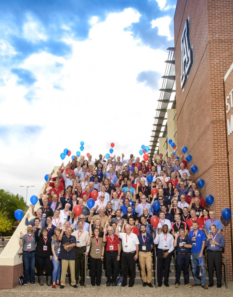 About 120 people pose for a photo on a large staircase outside the UA Student Union, flanked by red and blue balloons, and against a backdrop of a beautiful cloudy sky.