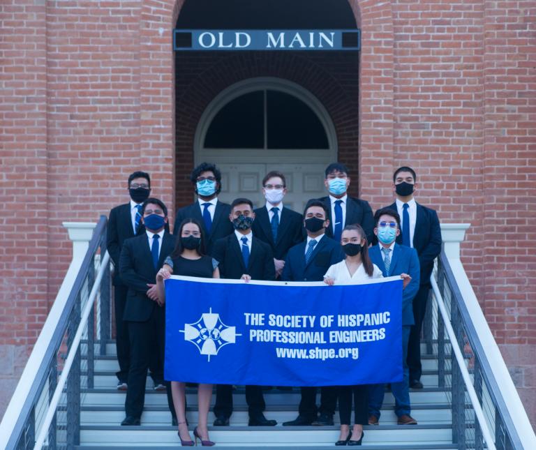 A group of students stands on the front steps of Old Main, wearing face masks and holding up a blue banner that reads "Society of Hispanic Professional Engineers. www.shpe.org."