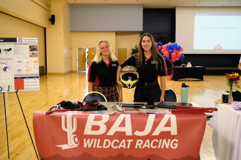 Two young woman stand behind a table with a red tablecloth that reads "Wildcat Baja Racing." One of the students holds a helmet under her arm.