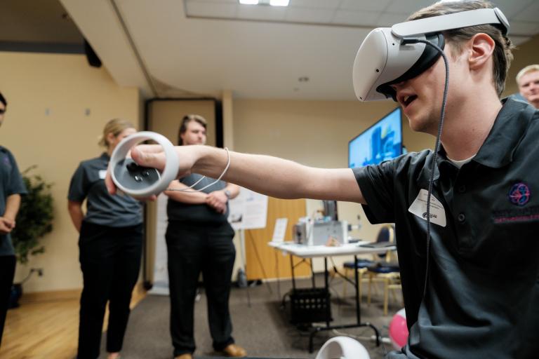 a student wearing a VR headset and holding a remote reaches out his arm with an expression of awe.