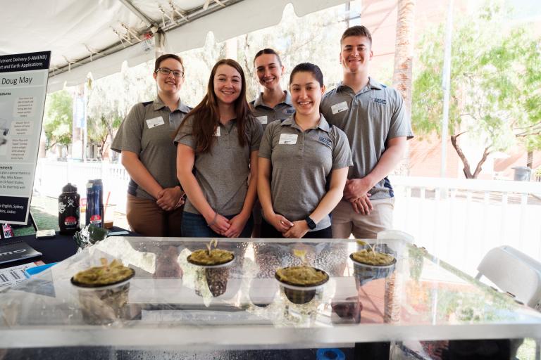 a group of five students stands behind a surface n which several plants are growing in small cups, like a muffin tin.