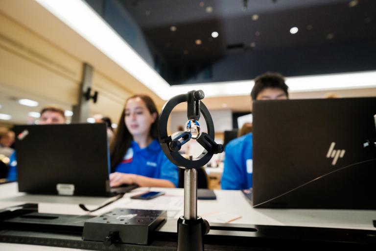 in the foreground, an optical device, with a concave lens held in a black 2D circular object balanced on a metal pole. Two students work on laptops in the background.