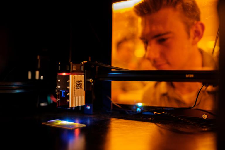 photo taken from inside an optical device.  a metal object shines a stream of light onto a chip. From the other side of an amber-colored glass, a student oversees the process.