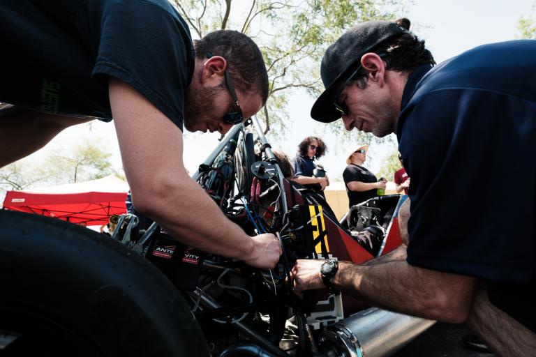 several students lean over a Formula 1 racing vehicle, while others look on from the background