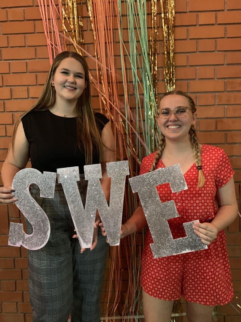 Two smiling women holding up cardboard cutouts of the letters "S W E"