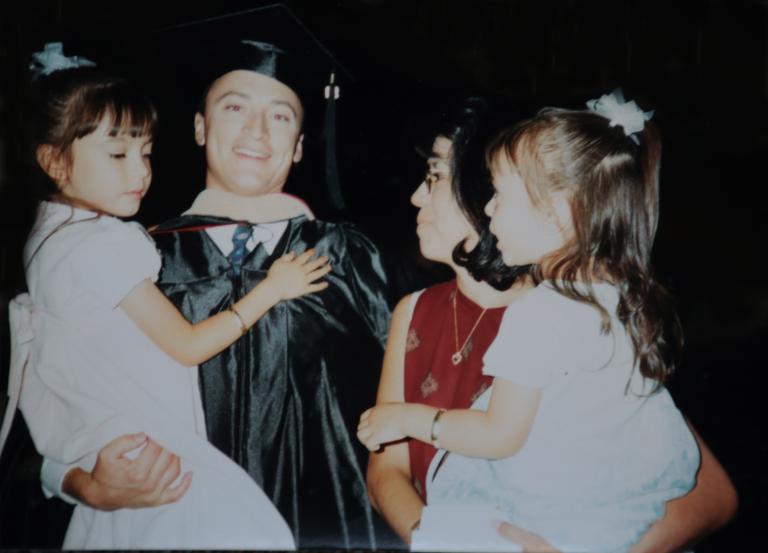 A man in a graduation cap and gown holds a young girl. Next to him is a woman in a red top holding another young girl.