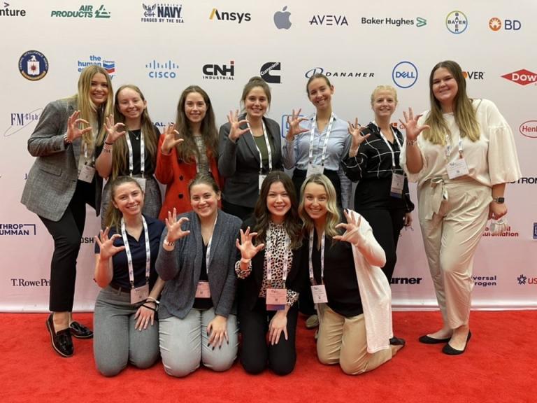 A group of young woman poses for a photo on a red carpet, in front of a backdrop.