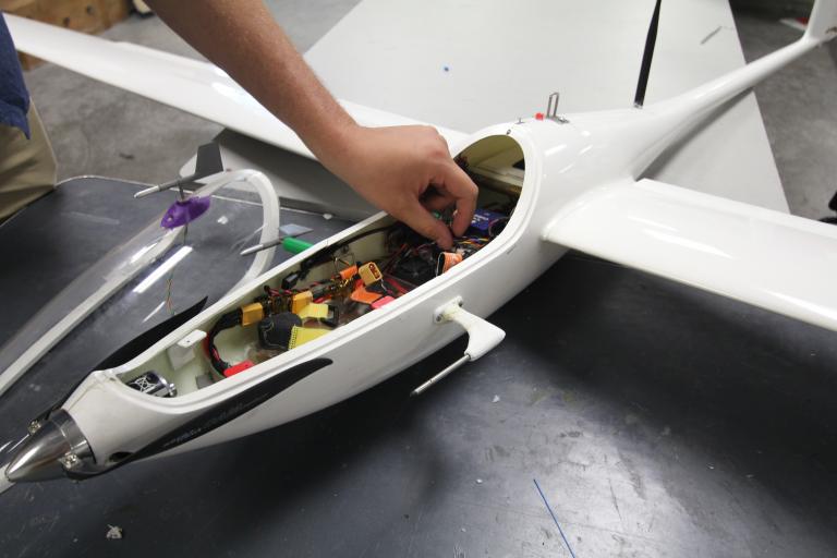 A closeup of the front part of a sailplane, with the transparent plastic cover taken off. A hand is adjusting one of the electronic components.