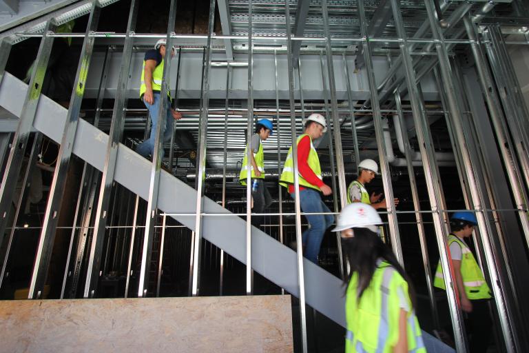 people in construction vests walk horizontally across the frame, up a set of stairs