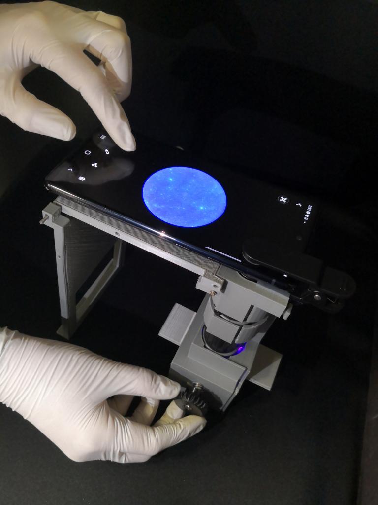 A smartphone with a blue circle on the screen sits on top of a boxy gray device. A pair of gloved hands are adjusting the phone.