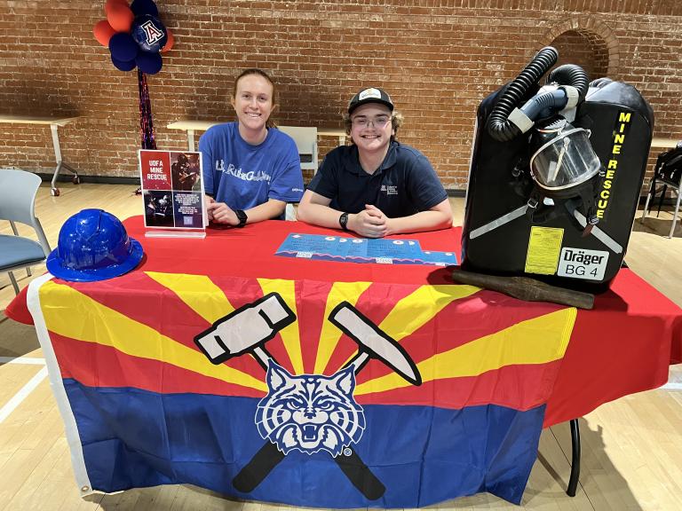 Two mining students smile at a student club event. They are pictured sitting behind a table with a mining hat to their right and a breathing apparatus to their left.