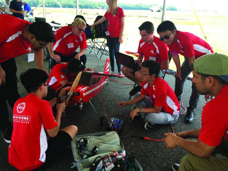 A group of students in red shirts huddle around an airplane, which is about 6 feet long.