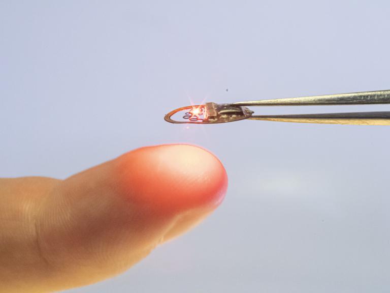 A fingertip enters the left side of the frame. Above it, held between a pair of tweezers, is a small, flat, metal, ring-shaped device with a red light on it. It is about half the size of a fingernail.