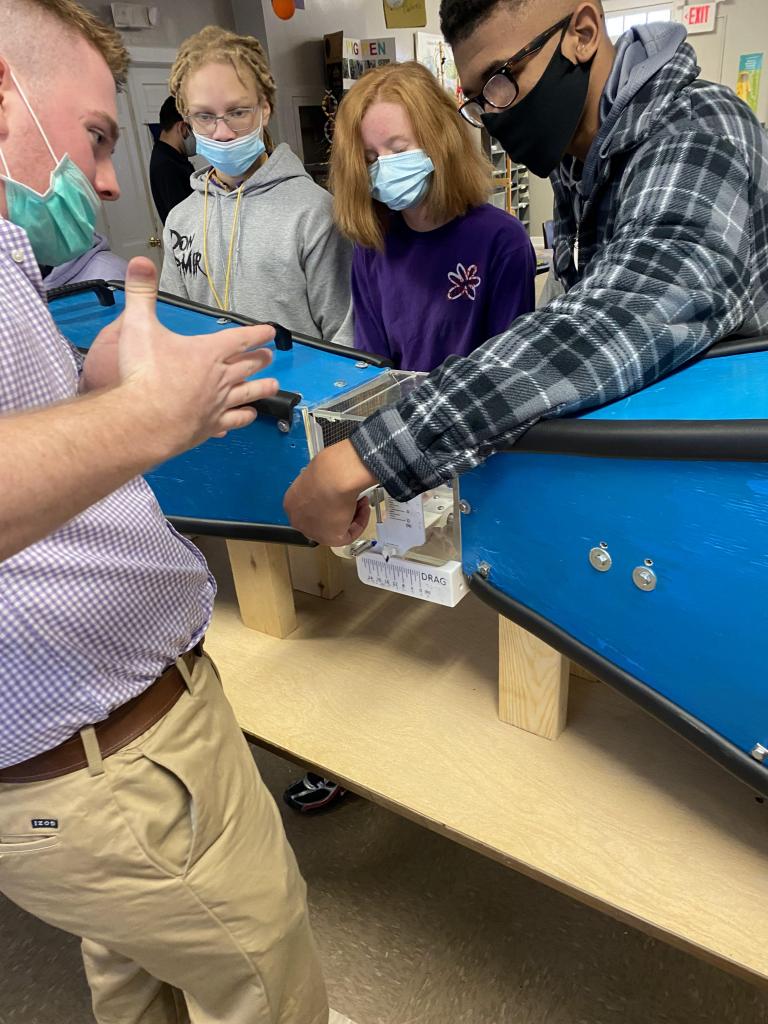 a group of four masked people leans over a small wind tunnel, which looks like two square blue funnels faced away from each other and connected by a transparent box.