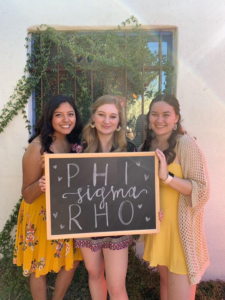 Three smiling women hold a blackboard with the words "Phi Sigma Rho"