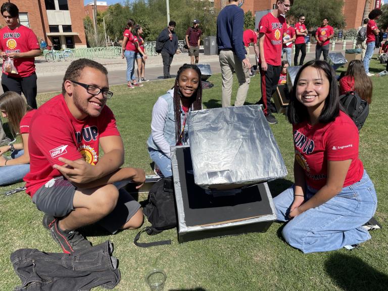 Three students surround a solar oven, a small cardboard box coated in foil.