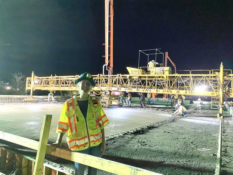 A young woman in a hard hat and yellow vest stands on a construction site and smiles. Other workers are pouring concrete behind her.