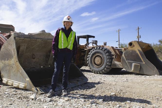 Liesl Folks, wearing a hard hat and a yellow vest, stands in front of mining equipment.