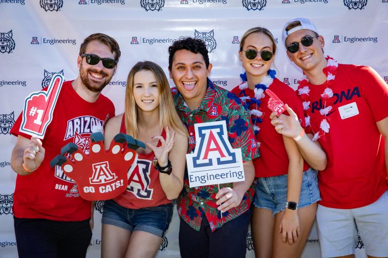 A group of five people stand in front of a UA Engineering backdrop holding props and decorations like leis, a foam finger and a Block A on a stick.