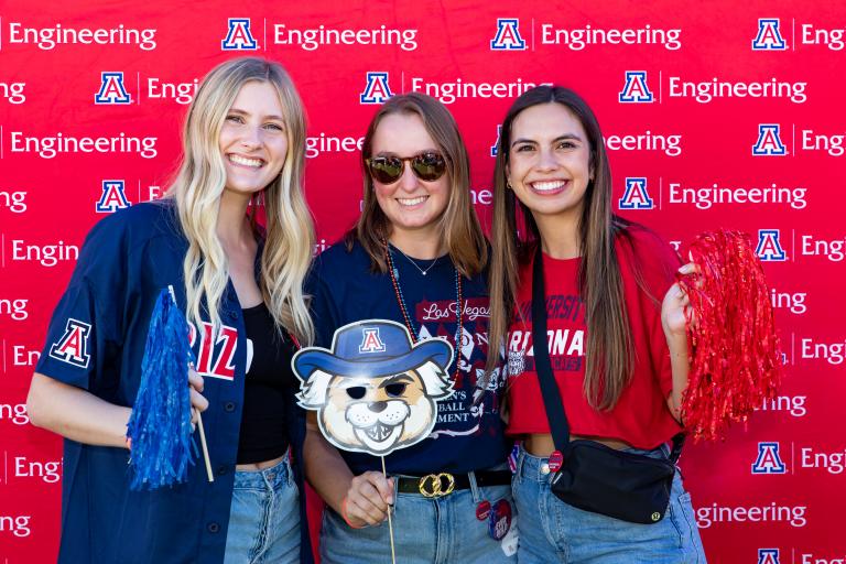 three young women stand in front of a red UA Engineering backdrop smiling and holding props, like a paper cutout of Wilbur Wildcat