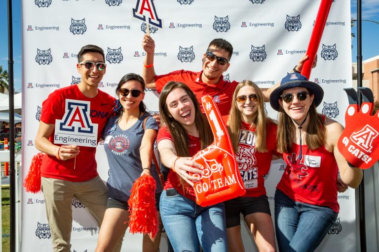 a group of students smile and hold up props like foam fingers in front of a white UA Engineering backdrop