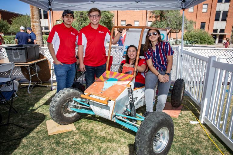 a group of students sit and stand around a Baja racing car on the UA Mall during the Engineering Homecoming tailgate