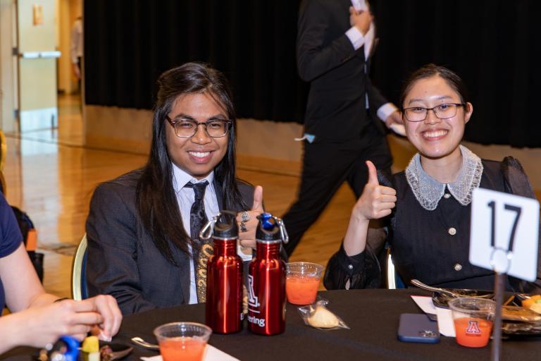 Two people sit at a formal table at an event, smiling and holding thumbs up.