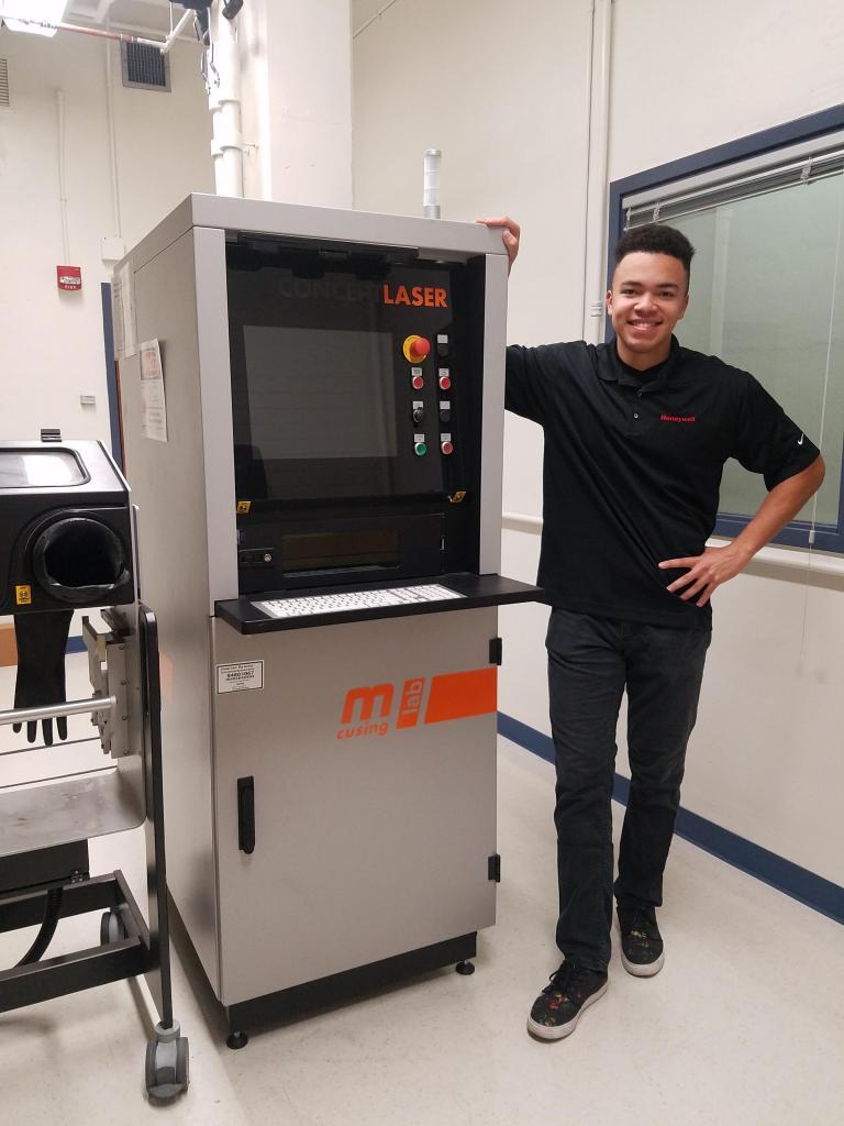 A man stands next to a 3D printing machine and smiles.