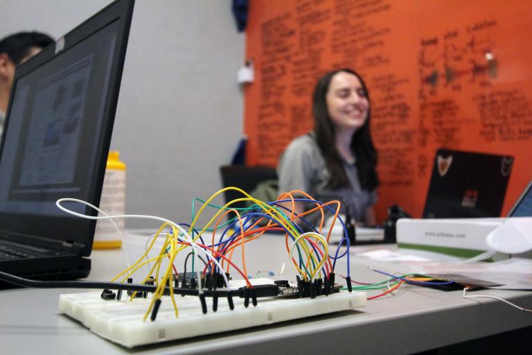 a board with colorful wires on it in the foreground. to the left is a laptop. in the background, out of focus, a young woman is smiling.