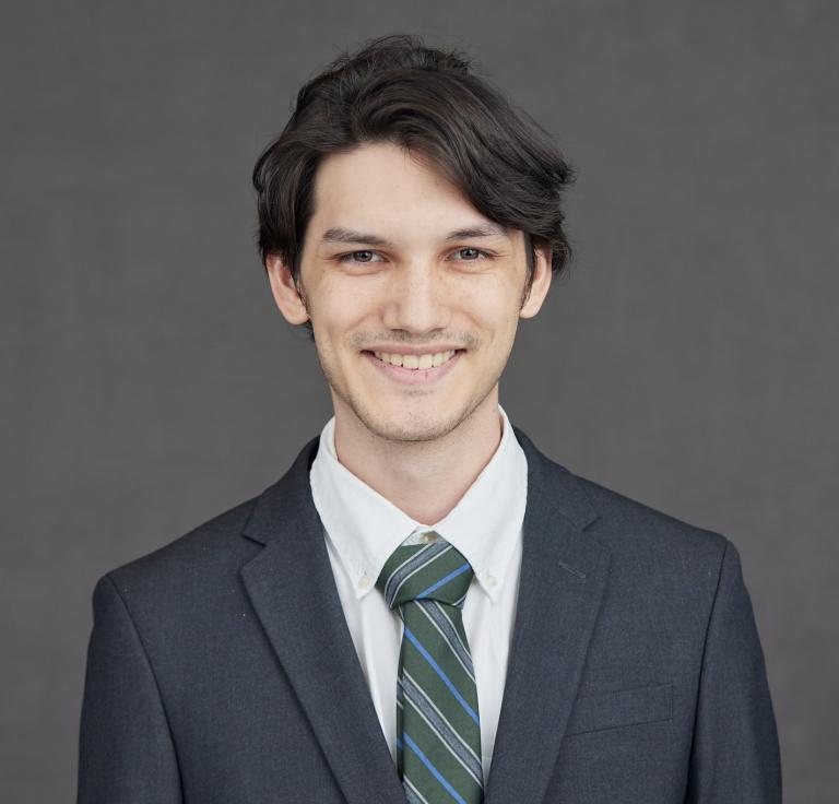 A man with brown hair wears a grey suit and tie.