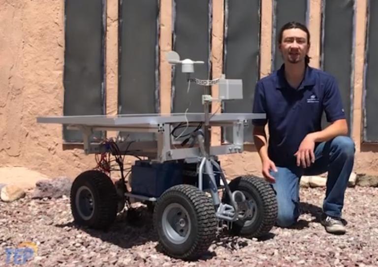 A man in a blue polo squats next to a rover, about the size of a toy jeep for a child. It has large wheels and there are solar panels on top.