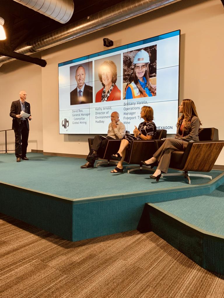 Three people sit in chairs on a stage, with a powerpoint with their photos behind them. One man stands and asks them questions.