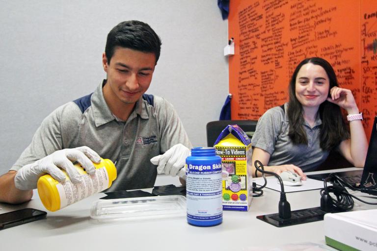 A young man wearing gloves pours a chemical into a tray while a young woman on a computer looks over at him from her laptop.