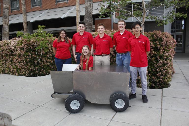 Five students stand around a big metal box on wheels. A sixth is sitting inside the box.