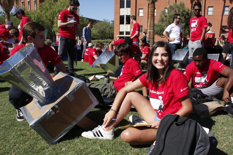A young woman sits next to a solar oven and smiles.