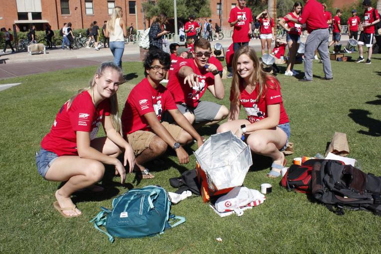 Four students in matching red shirts squat and pose next to a solar oven, a cardboard device about a foot across with a foil funnel-like structure on top.
